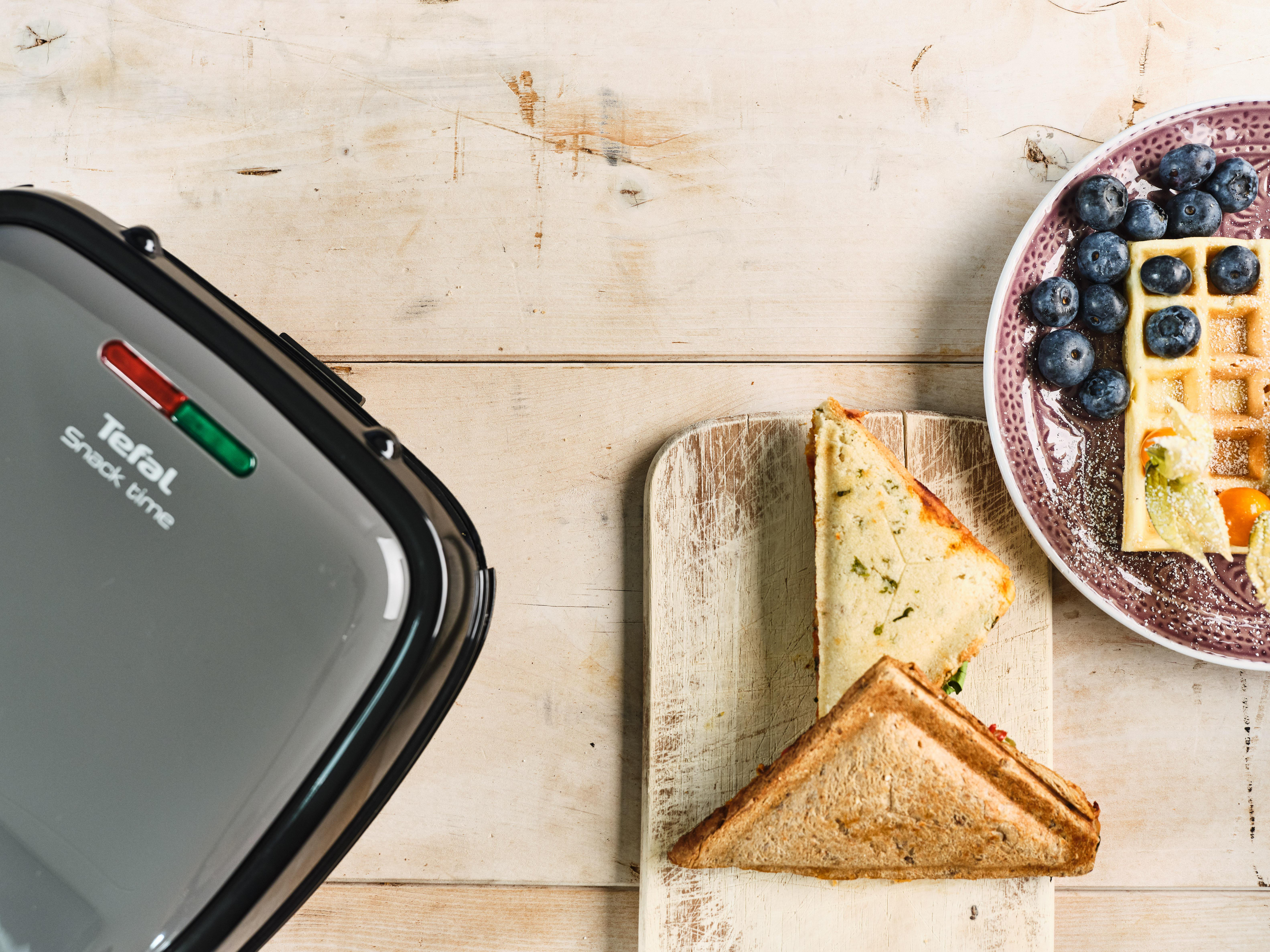 Waffeleisen mit Waffeln, einem Sandwich und einem Teller mit Blaubeeren.