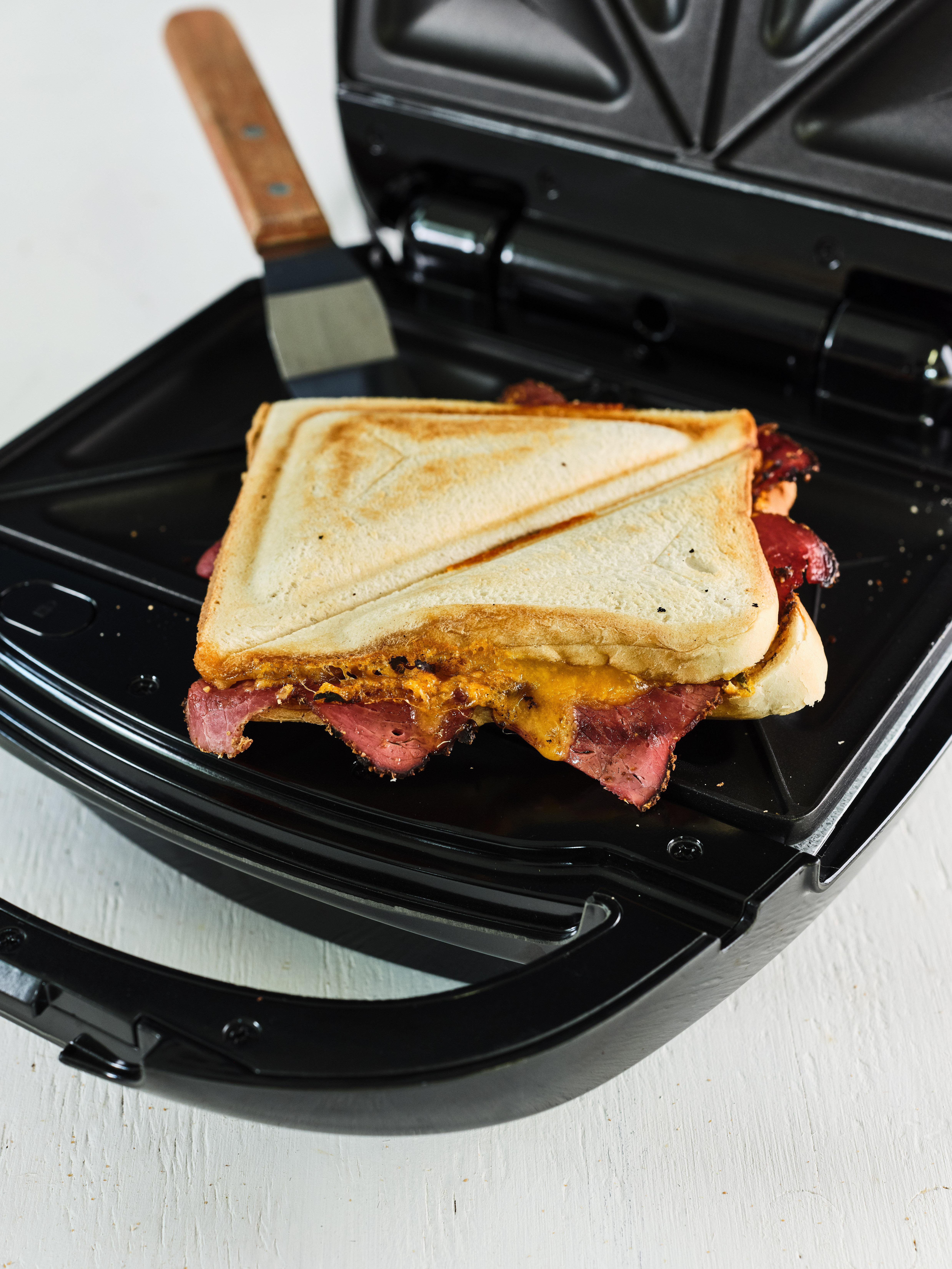 Sandwich in einem Sandwichmaker. Brot, Käse und Fleisch sind sichtbar. Der Maker ist schwarz.