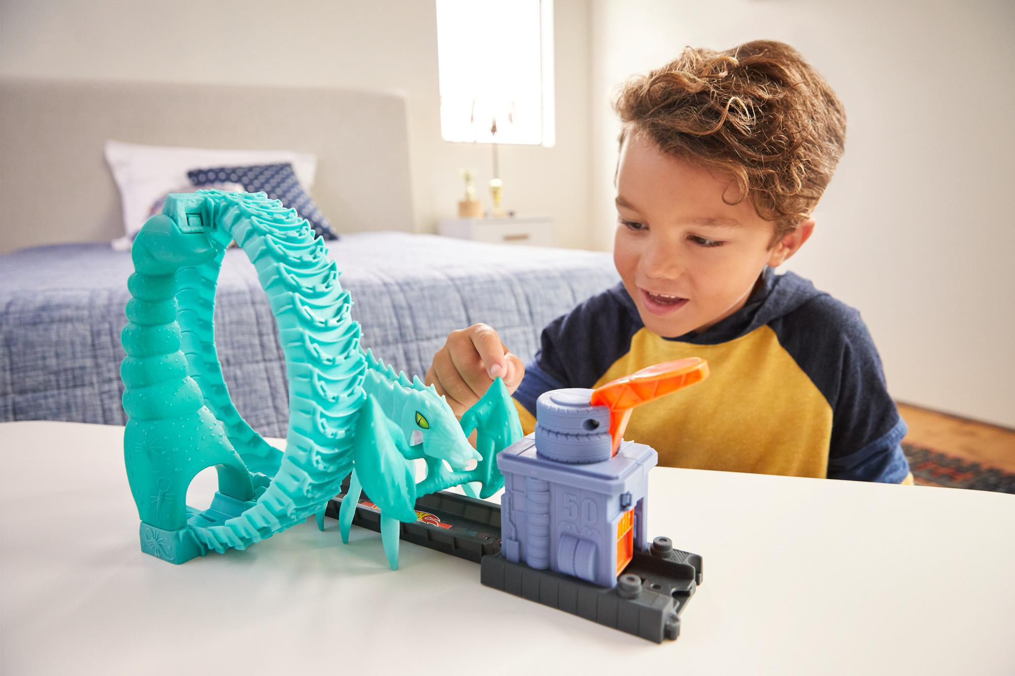 Ein Junge spielt mit einer Spielzeug-Autorennbahn. Die Bahn hat eine Schleife und einen Startmechanismus. Schlafzimmer und Bett im Hintergrund.
