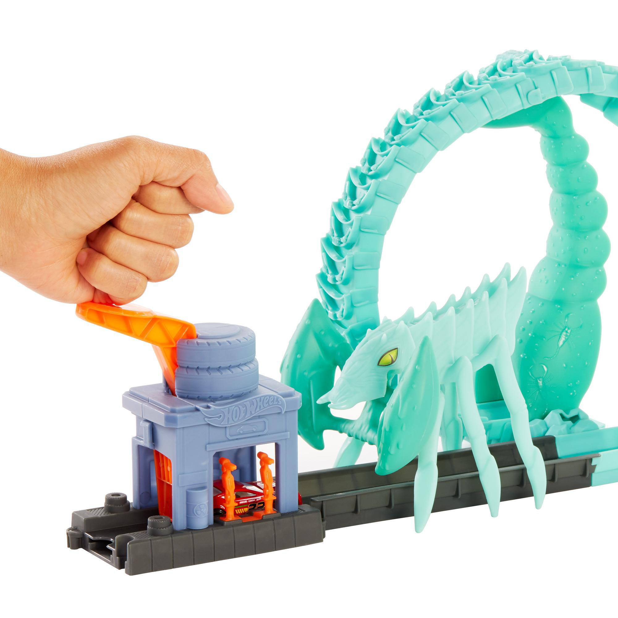 Eine Hand betätigt einen orangefarbenen Hebel auf einer Hot Wheels-Autorennbahn. Eine skorpionartige Struktur und ein rotes Auto sind sichtbar.