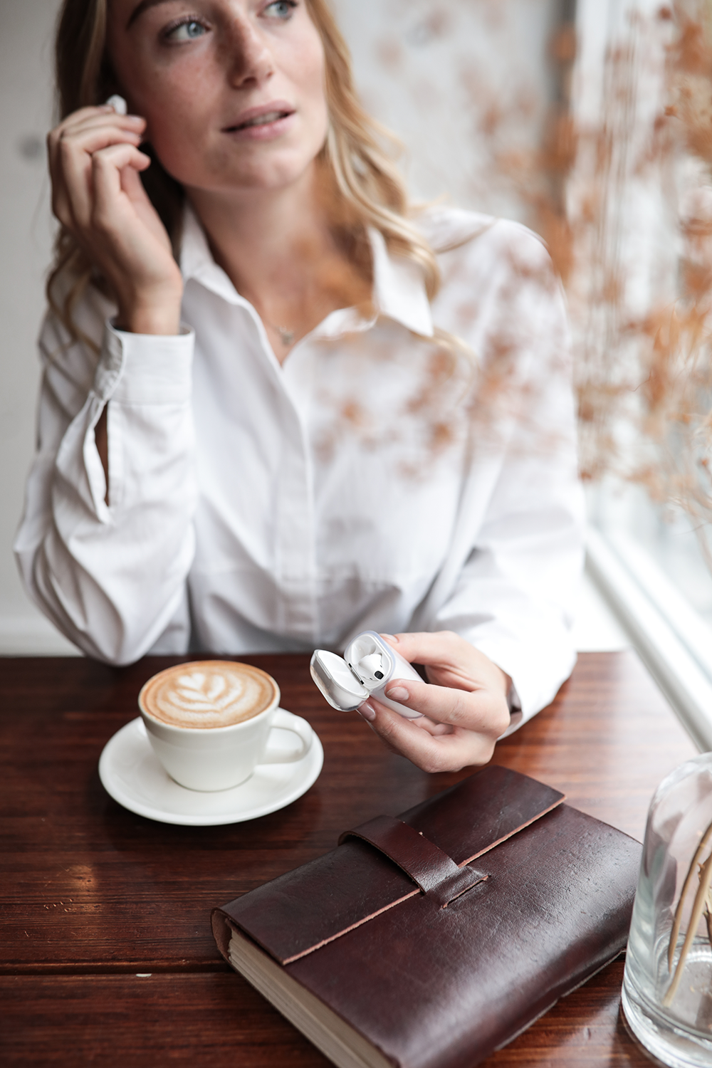 Woman in a white shirt holds earbuds and a case. Coffee and a notebook are on the table. 
