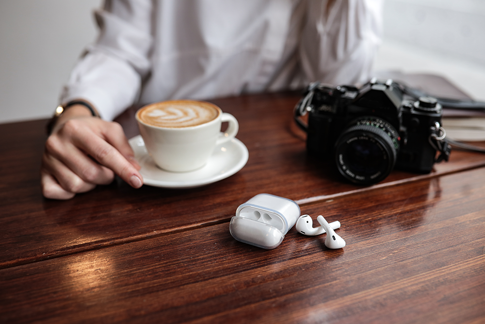 Coffee, camera, and AirPods on a wooden table. A person's hand is in the frame.
