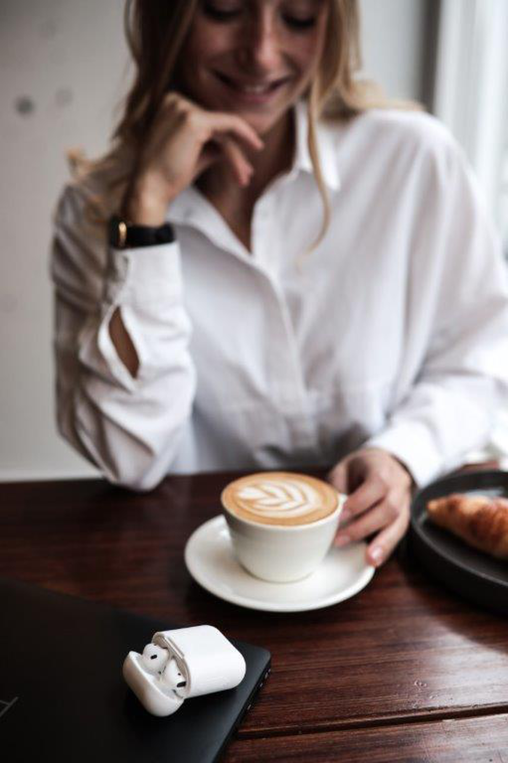 Vrouw met koffie en croissant. AirPods op tafel, laptop zichtbaar. Houten tafel.