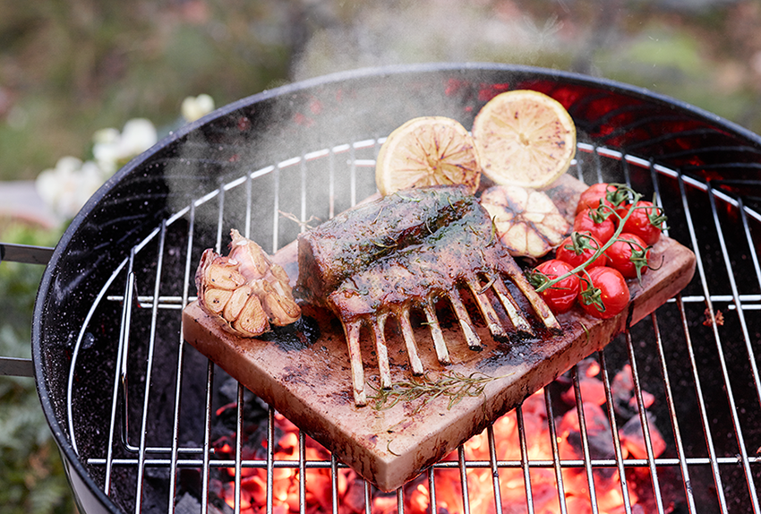 Grilled lamb rack on a wooden board, with tomatoes and lemon. On a BBQ grill with glowing coals.
