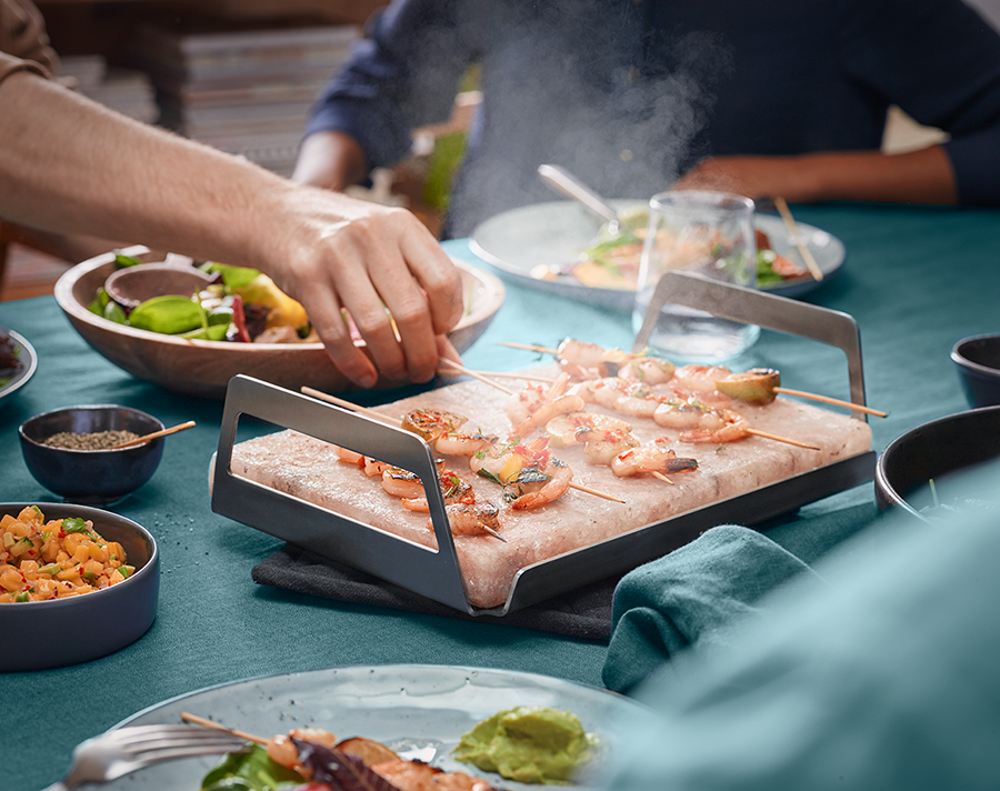 People grilling shrimp and vegetables on a salt block, with smoke rising from the food.