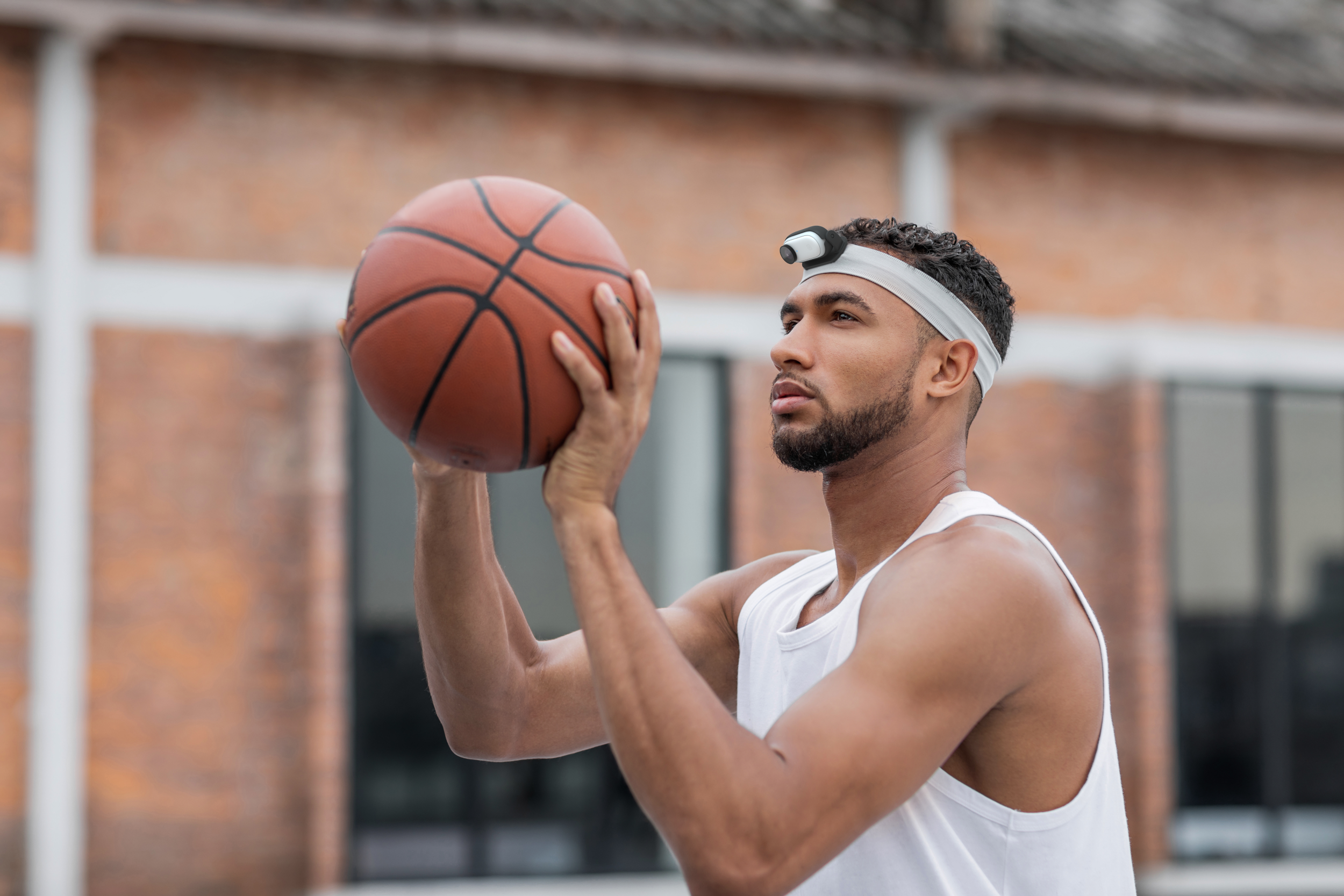 Ein Mann in einem weißen Tanktop, der einen Basketball hält und kurz davor steht, zu werfen, vor einer Backsteinmauer.