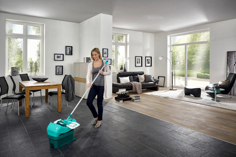 Frau putzt mit einem grünen Mopp den Boden in einem modernen Wohnzimmer.