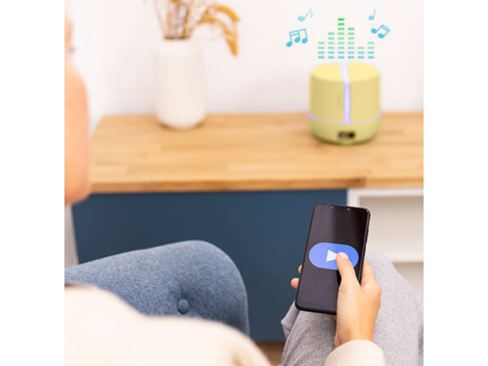 Una persona usa un teléfono con un botón de reproducción, cerca de un altavoz y notas musicales. En el fondo, una mesa de madera y un jarrón son visibles.