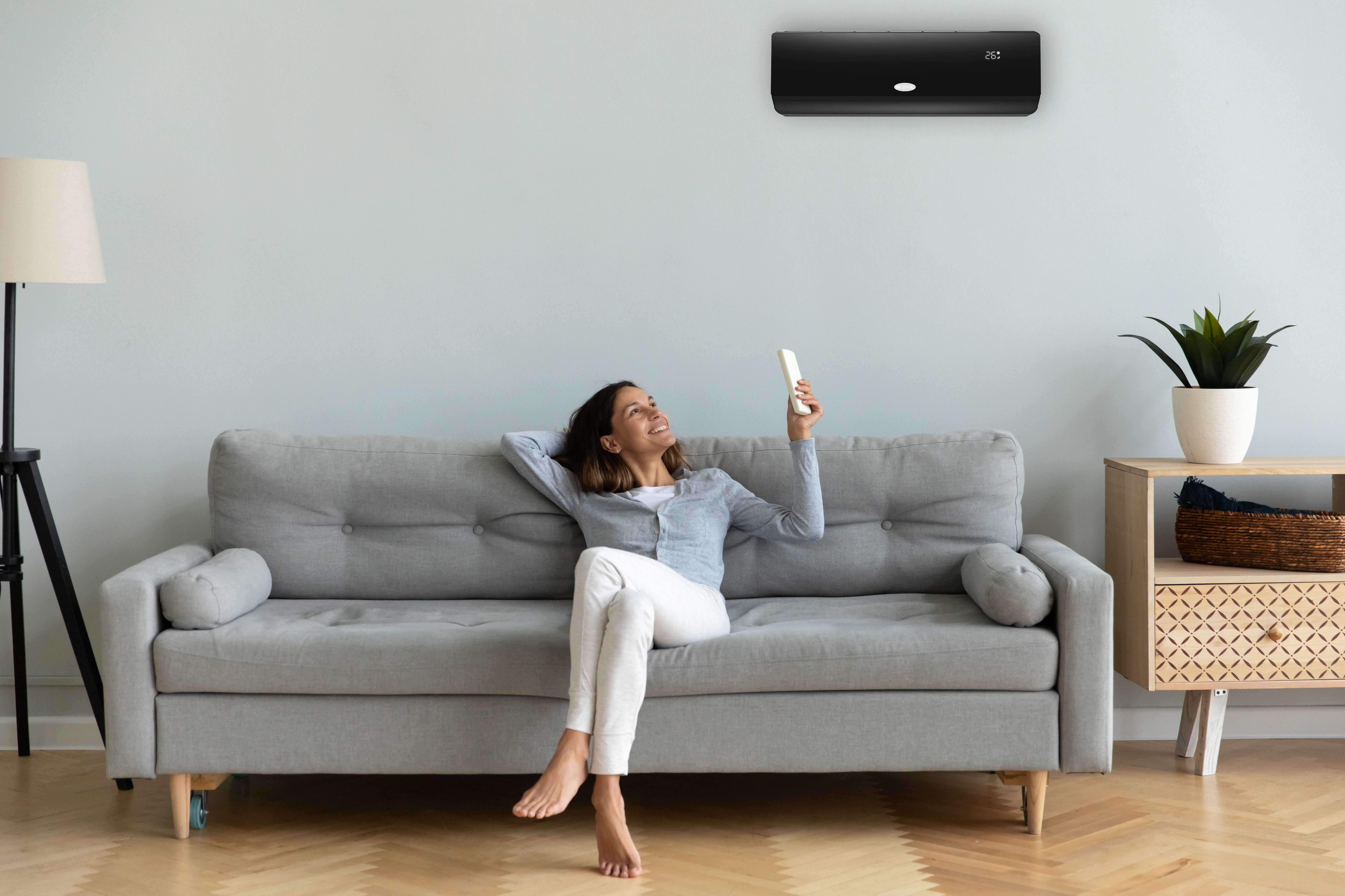Eine Frau sitzt auf einem Sofa und blickt auf eine schwarze Klimaanlage. Es gibt eine Lampe und eine Pflanze.