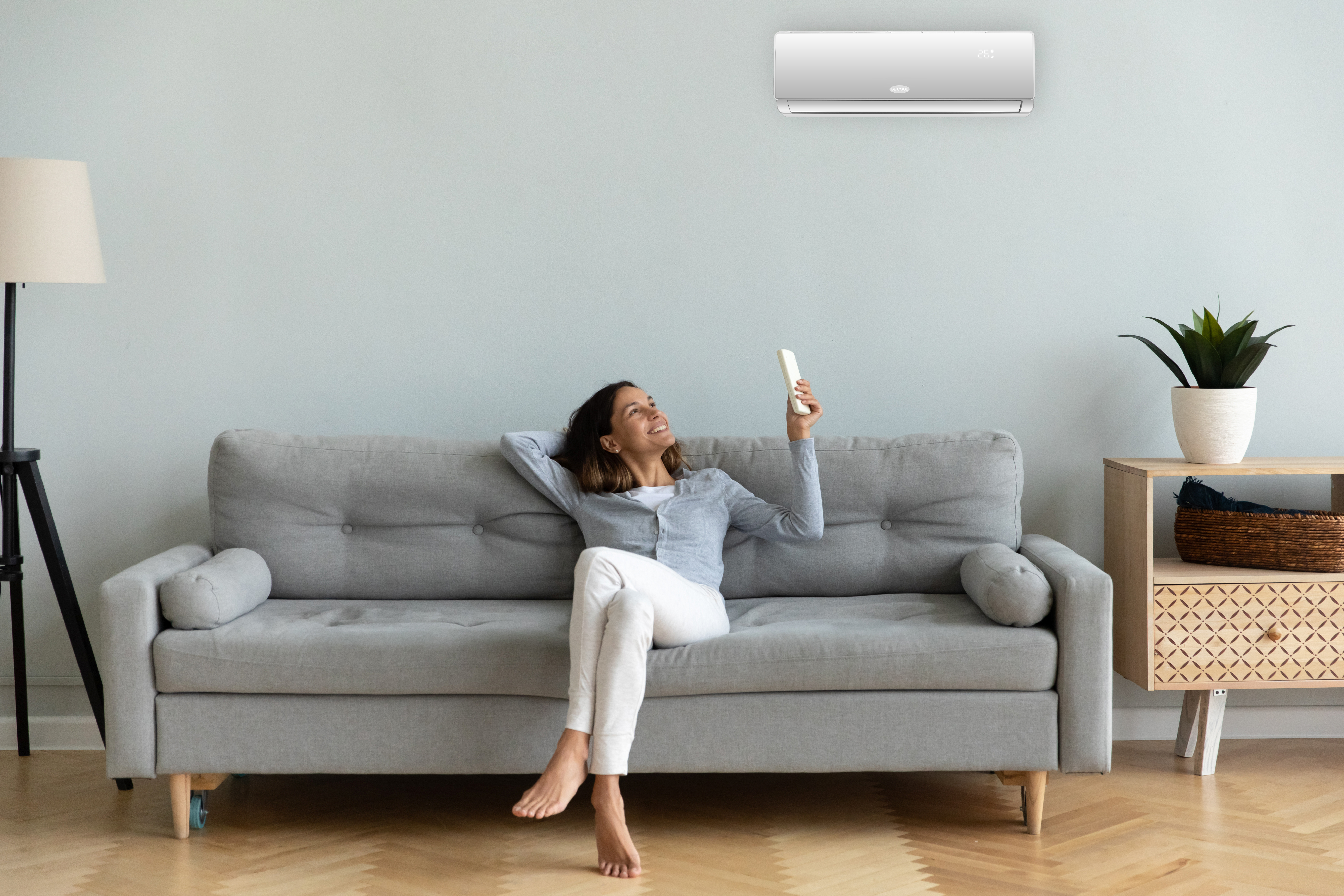 Frau auf grauer Couch, hält Fernbedienung, blickt auf Klimaanlage. Entspannende Szene.
