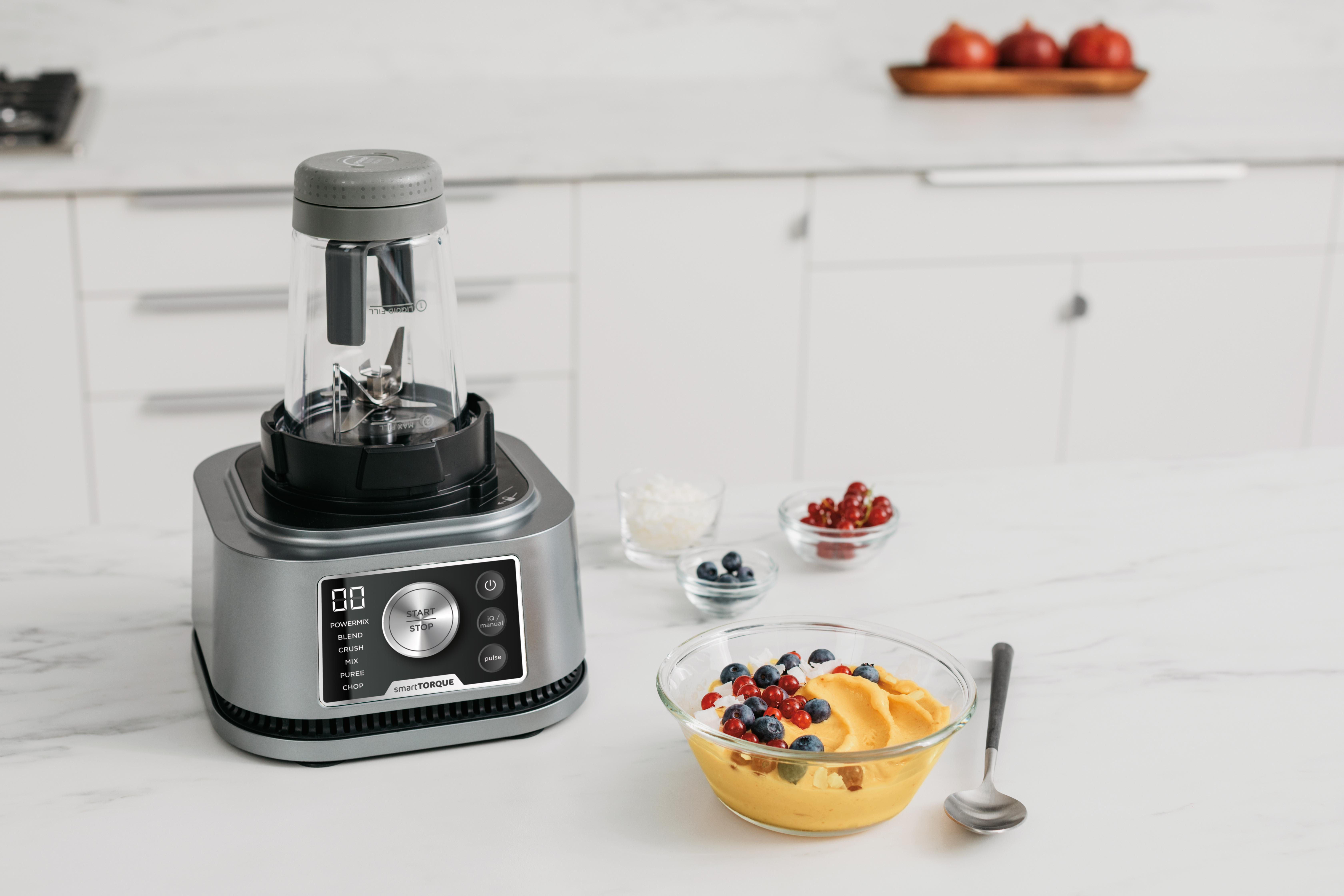 Ein Mixer mit einer Tasse Obst auf einer weißen Theke und Obstschalen in der Nähe.