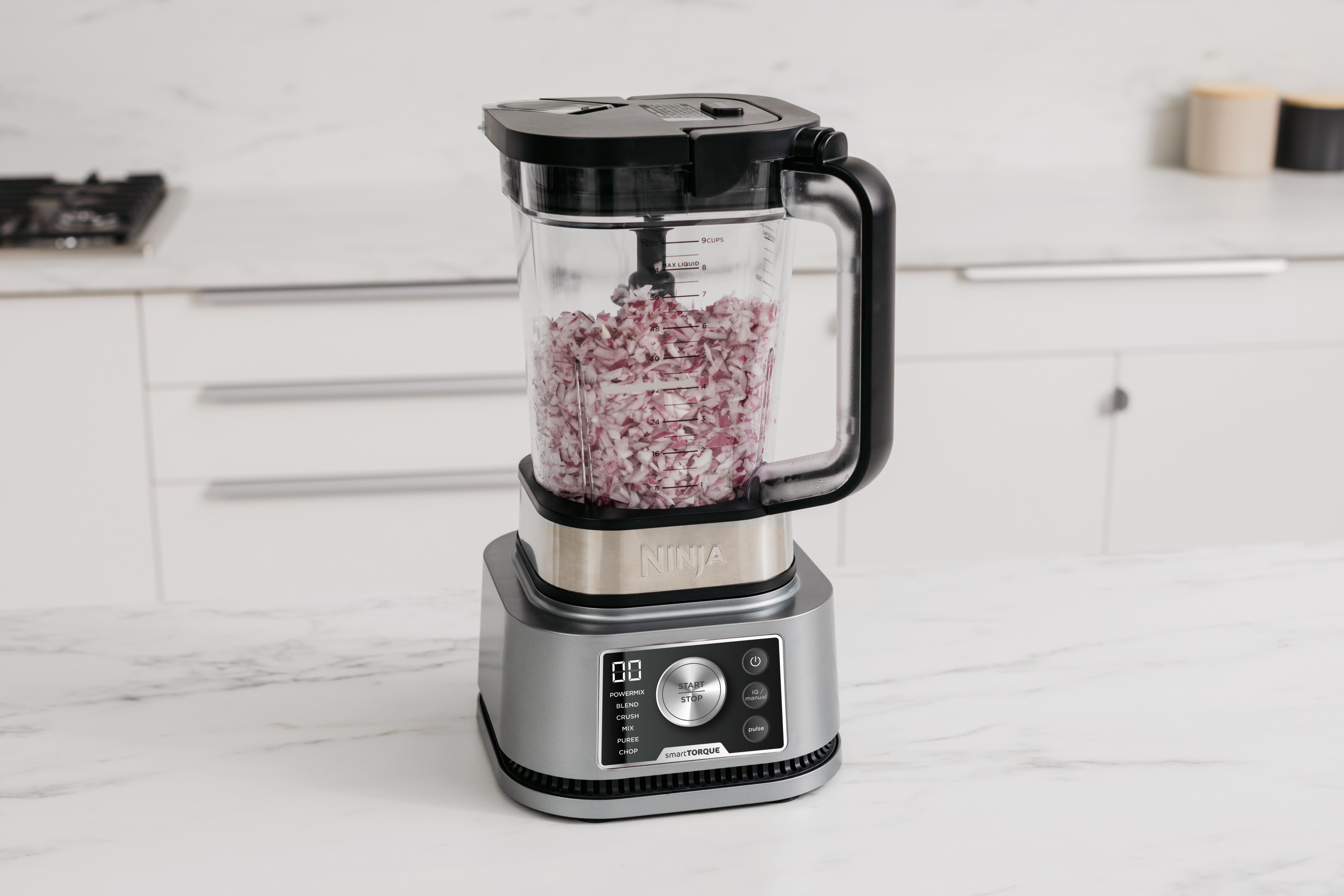 Ein Ninja-Mixer mit gewürfelten roten Zwiebeln im Inneren, auf einer weißen Arbeitsplatte.