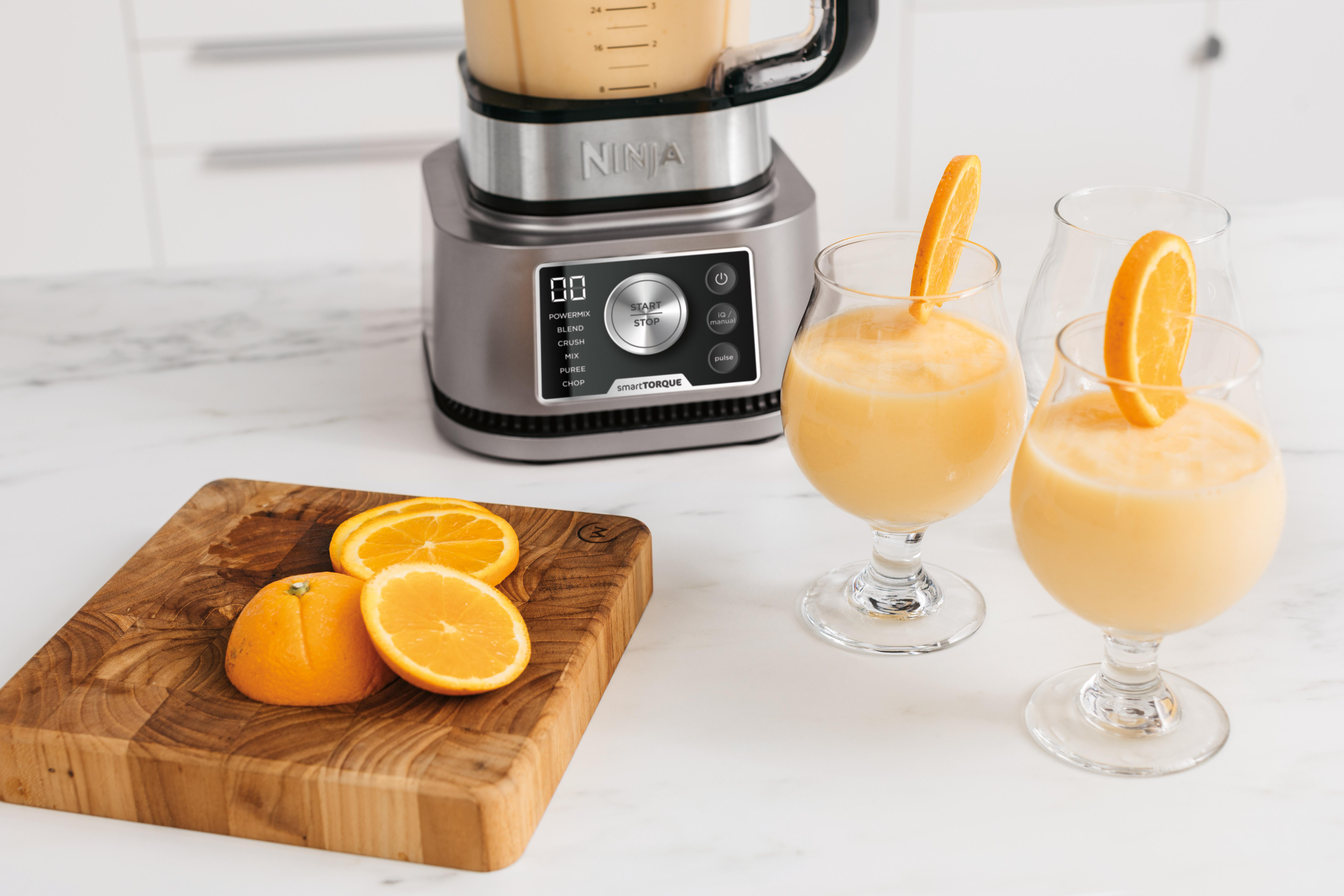 Ein Mixer mit Orangensmoothie und Orangenscheiben auf einem hölzernen Schneidebrett. Zwei Gläser mit Smoothie.