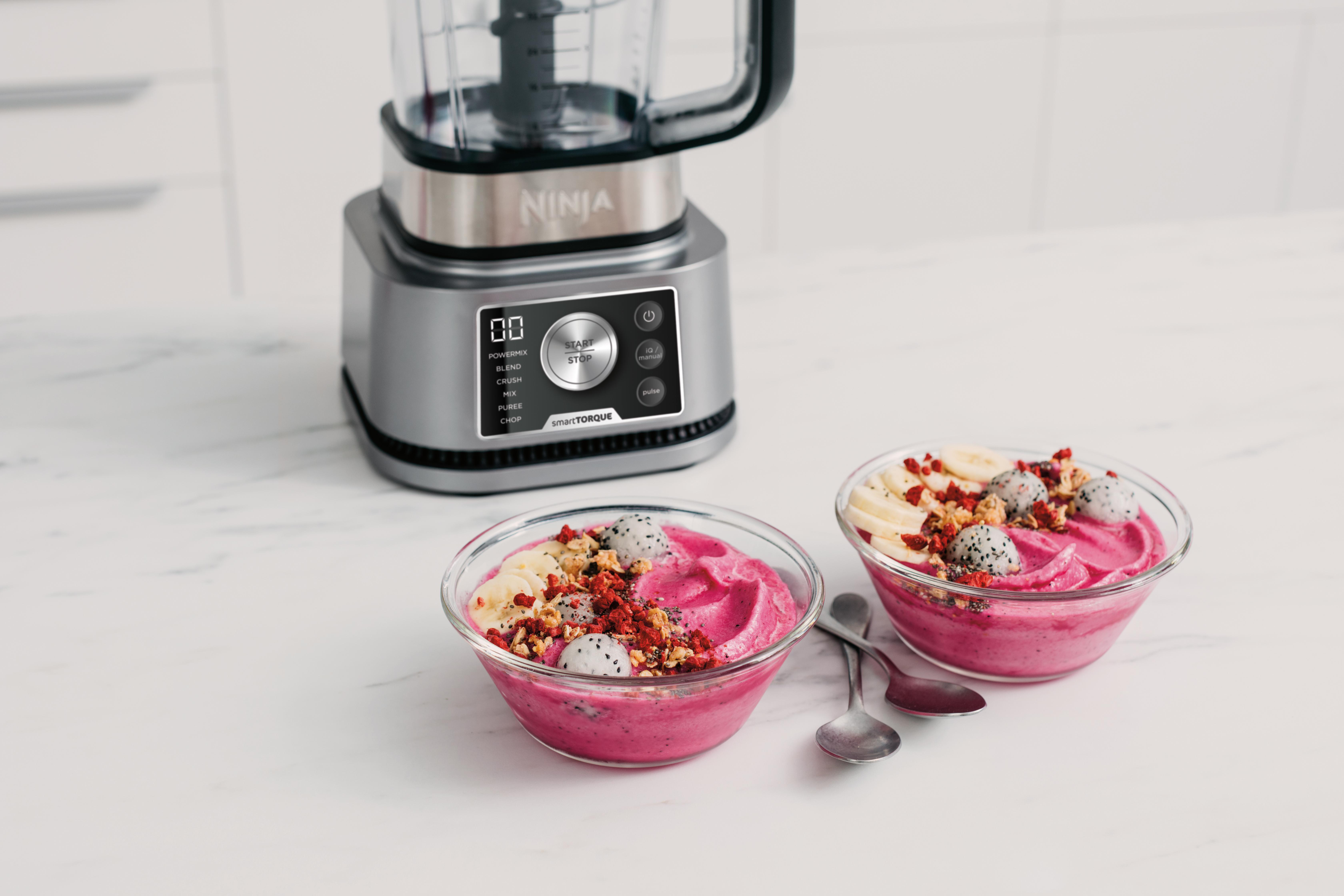 Zwei Smoothie-Schalen mit Toppings und ein Mixer auf einer weißen Marmor-Arbeitsplatte.