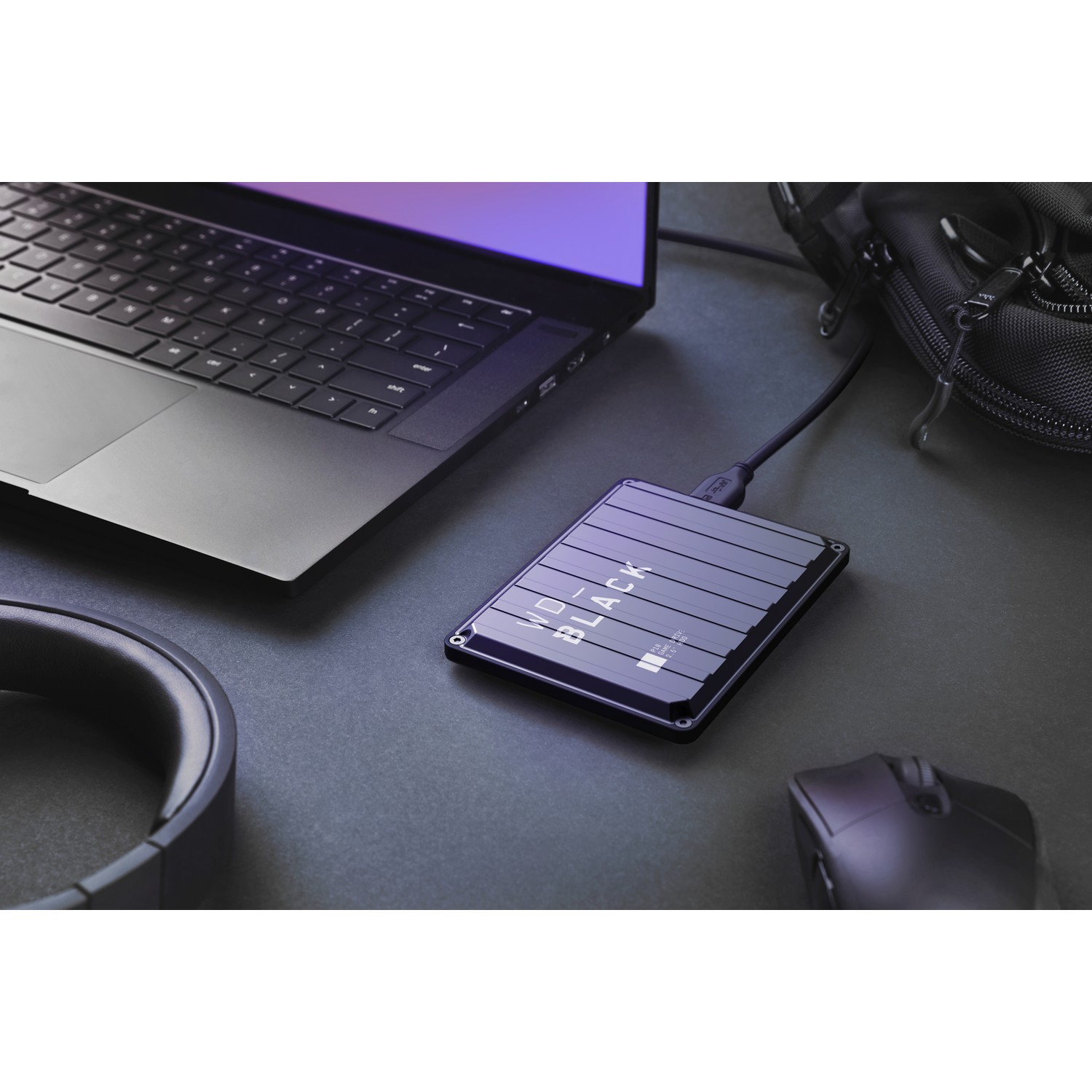 Un disco duro externo negro junto a un portátil, un ratón y auriculares sobre una superficie oscura.