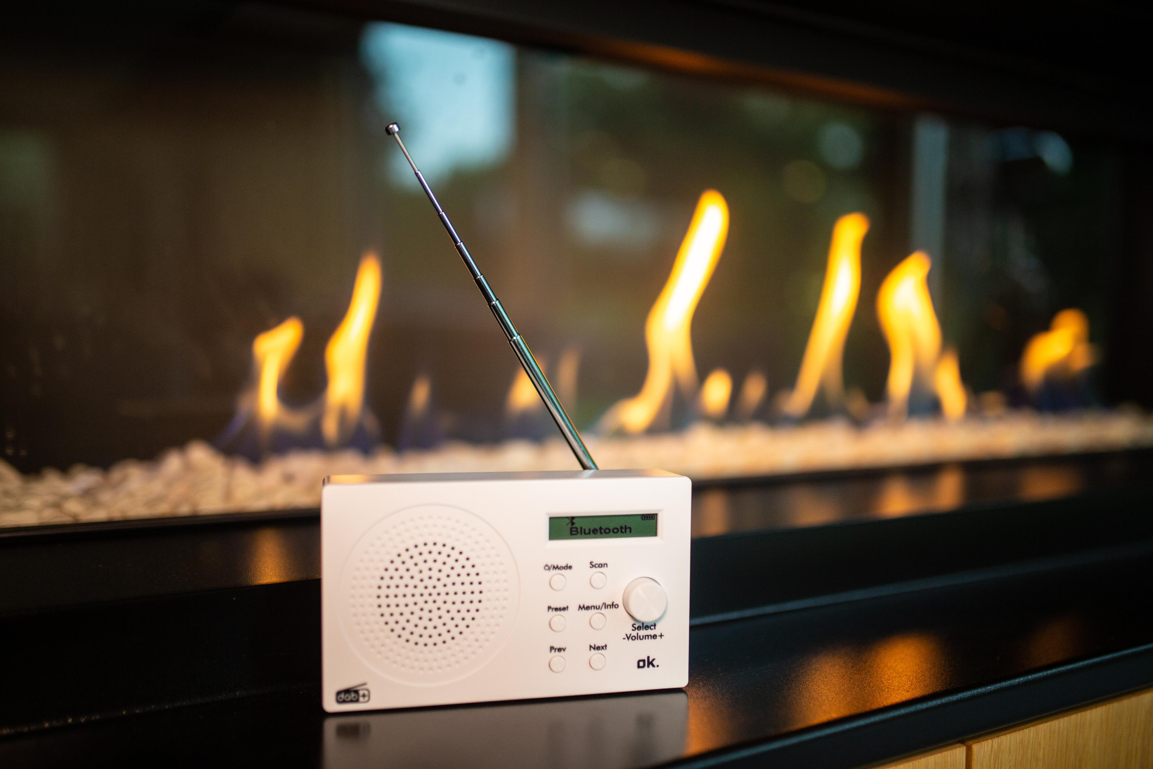 Weißes Radio auf schwarzer Oberfläche, Flammenhintergrund. Display zeigt Bluetooth und andere Informationen.