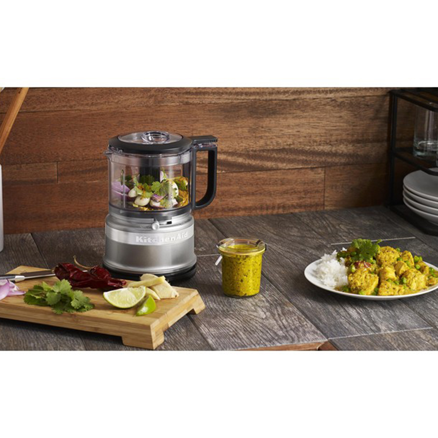 Scena cucina: robot da cucina con ingredienti, piatto di cibo e barattolo di salsa su superficie legno.