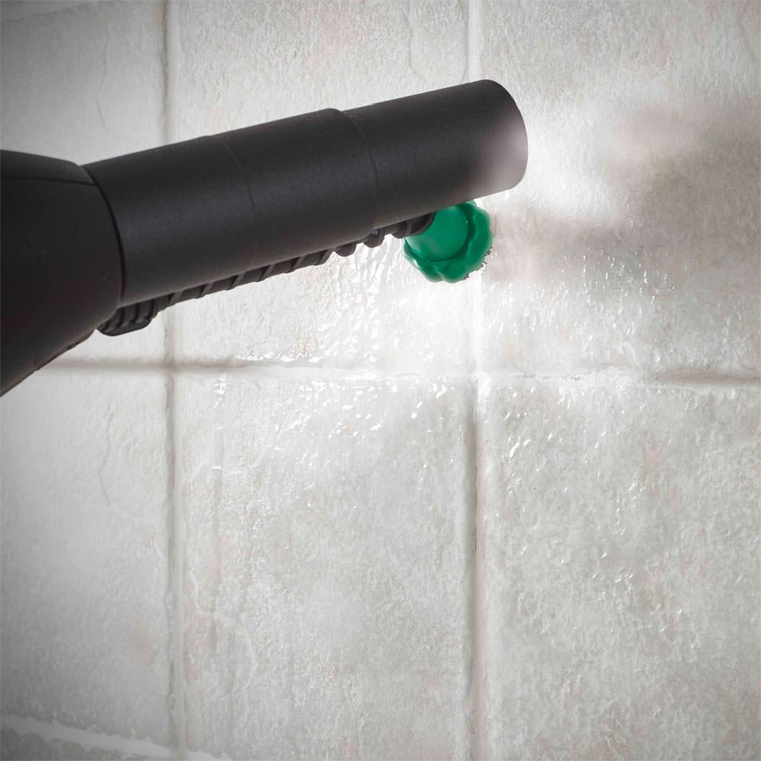 Primo piano di un ugello di un pulitore a vapore che spruzza acqua su un muro piastrellato bianco.