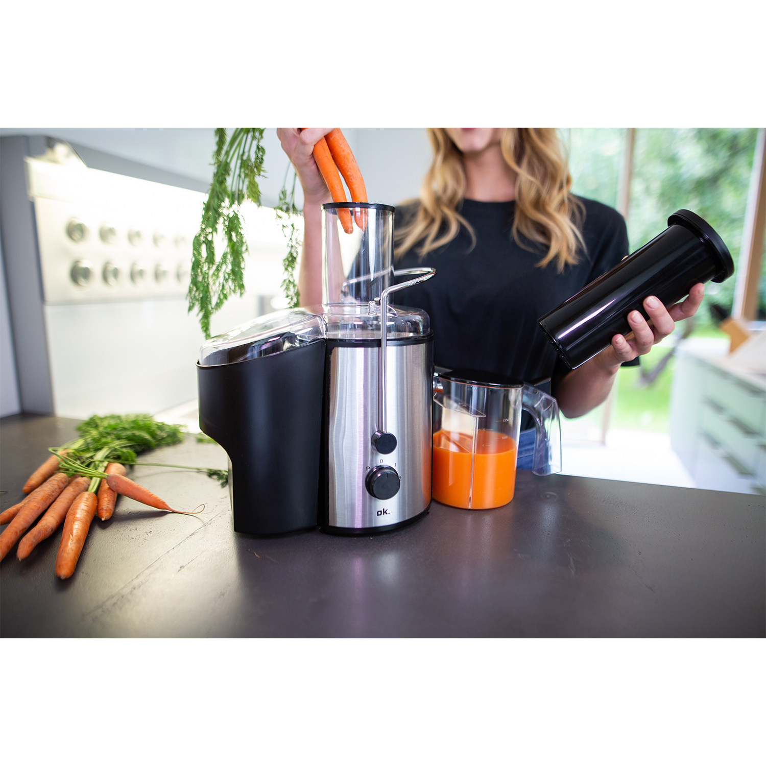 Una donna che usa un estrattore di succo, tiene carote, con un bicchiere di succo e un contenitore.