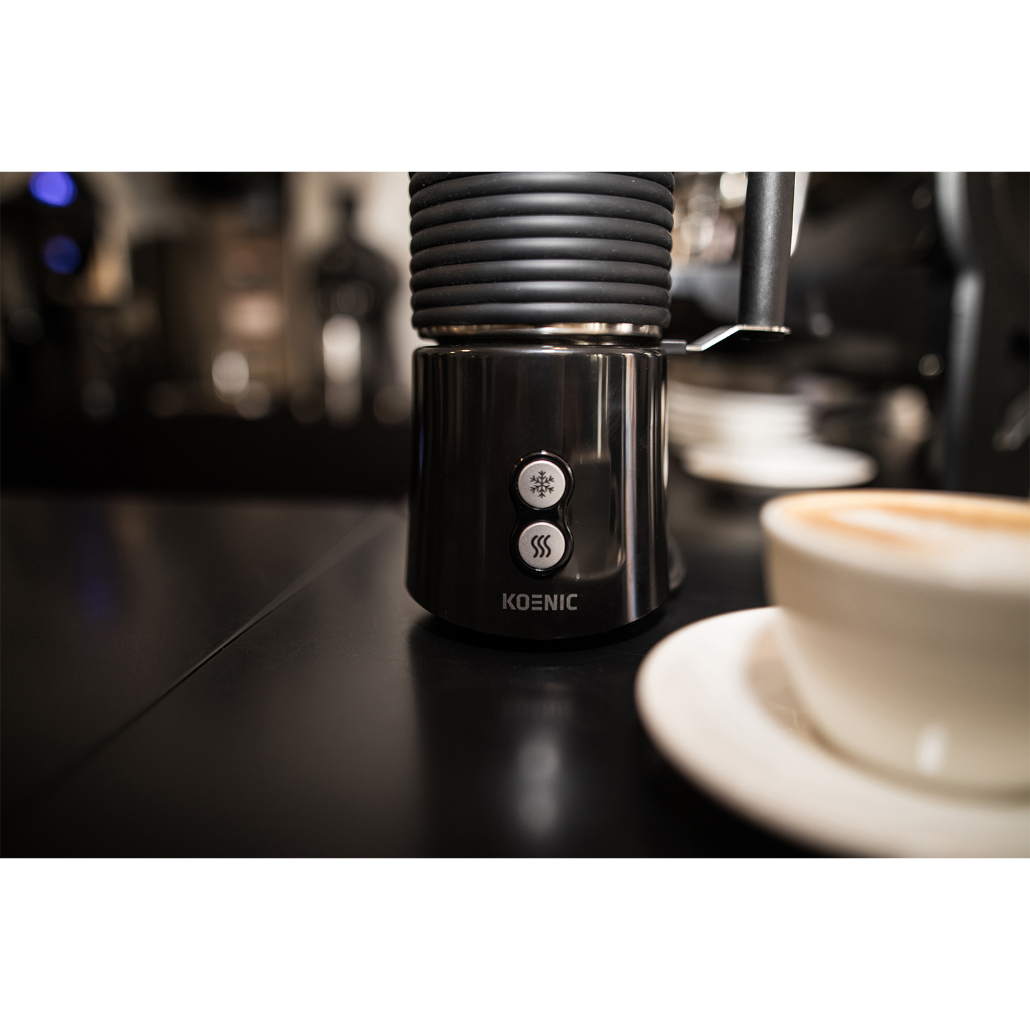 Montalatte Koenic nero con pulsanti, una tazza di caffè e sfondo sfocato.
