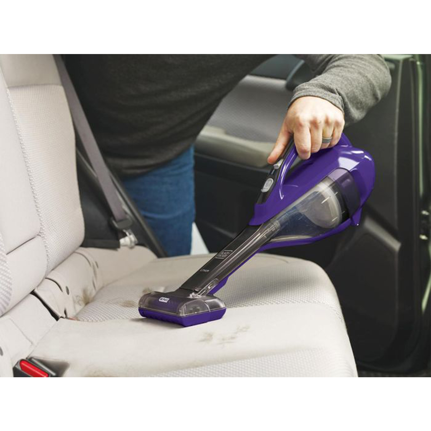 Una persona che aspira un sedile dell'auto con un aspirapolvere portatile viola. Il sedile è beige e macchiato.