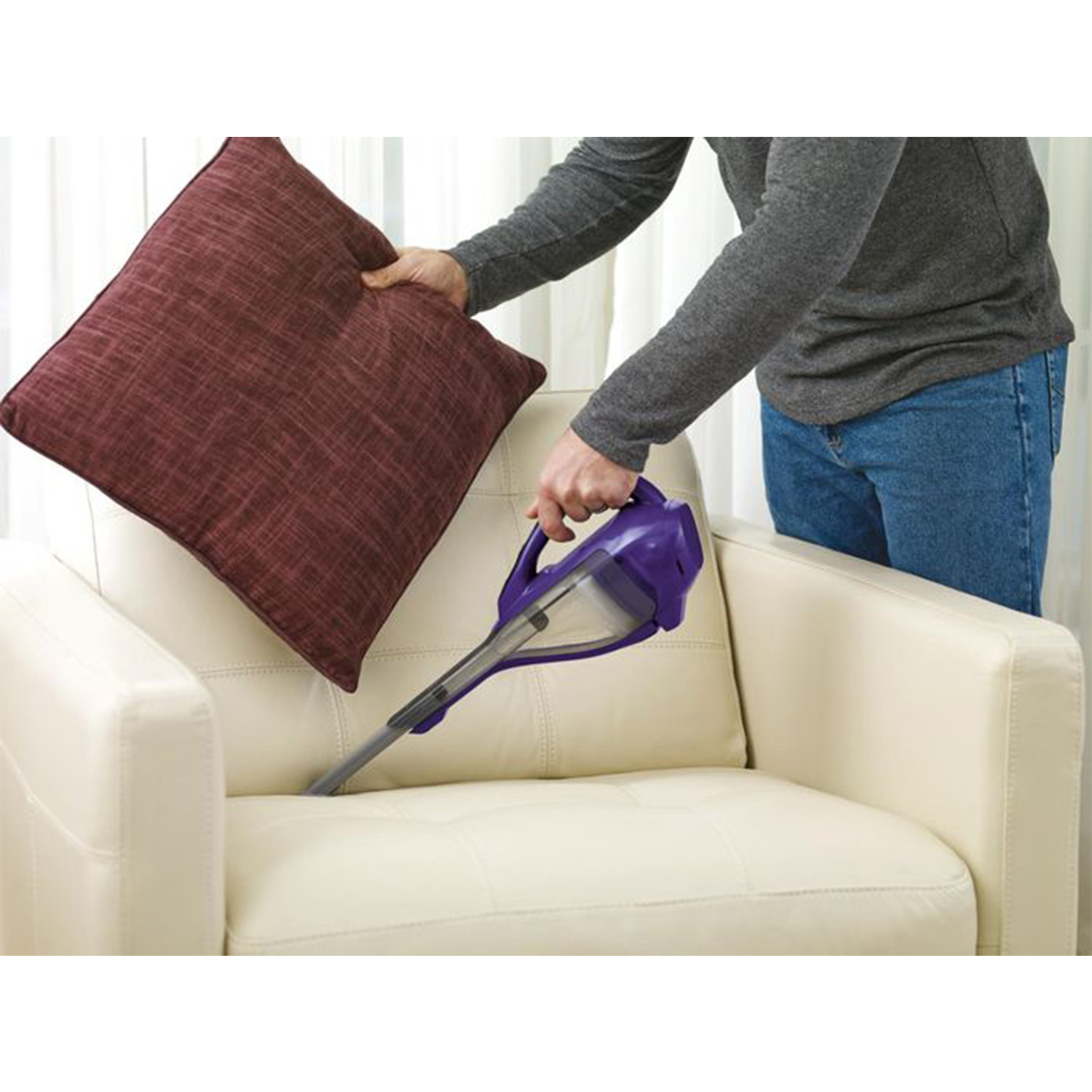 Una persona che aspira un divano color crema con un aspirapolvere portatile viola. Un cuscino bordeaux è accanto.