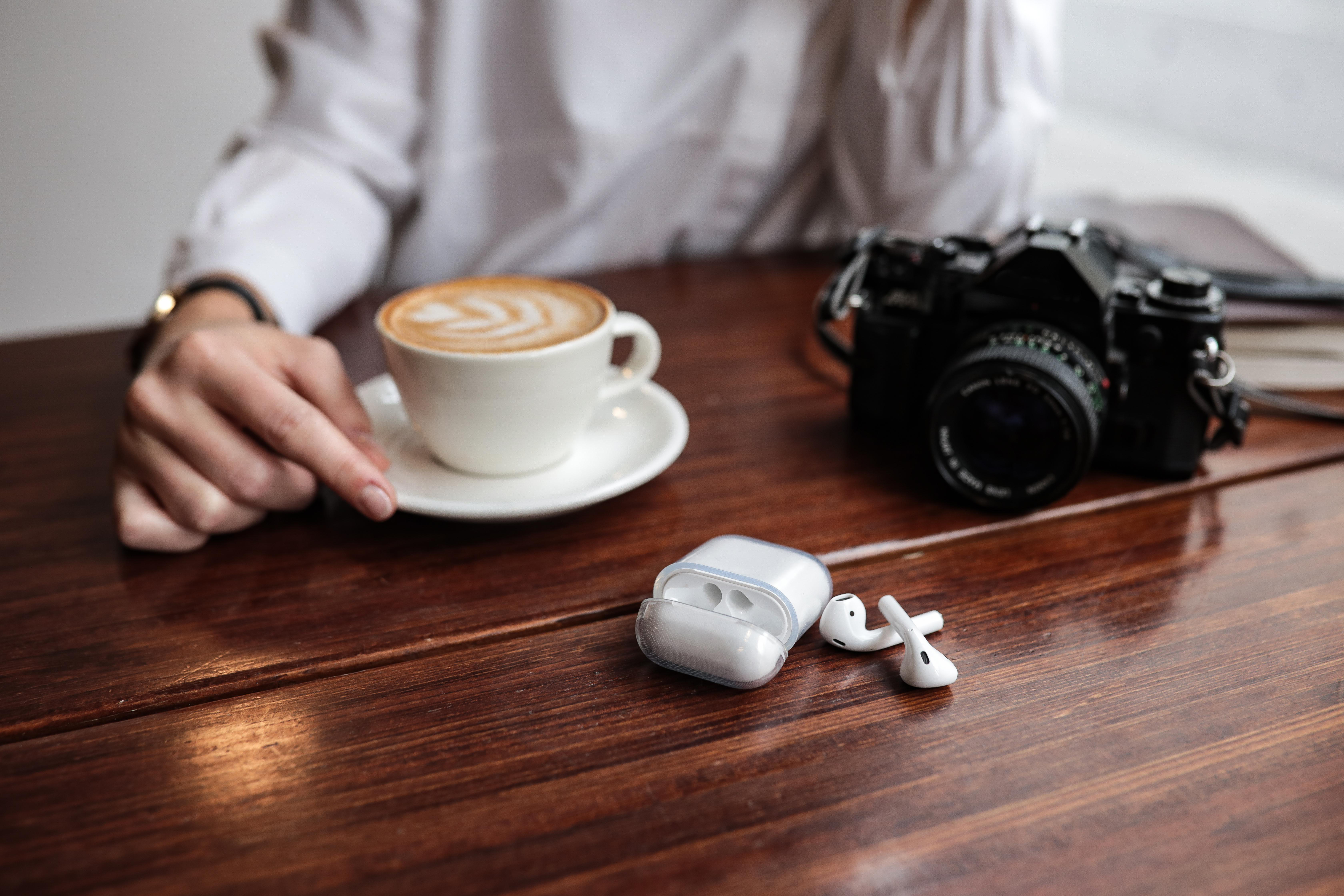 Person mit Kaffee, Kamera und Ohrhörern auf einem Holztisch.