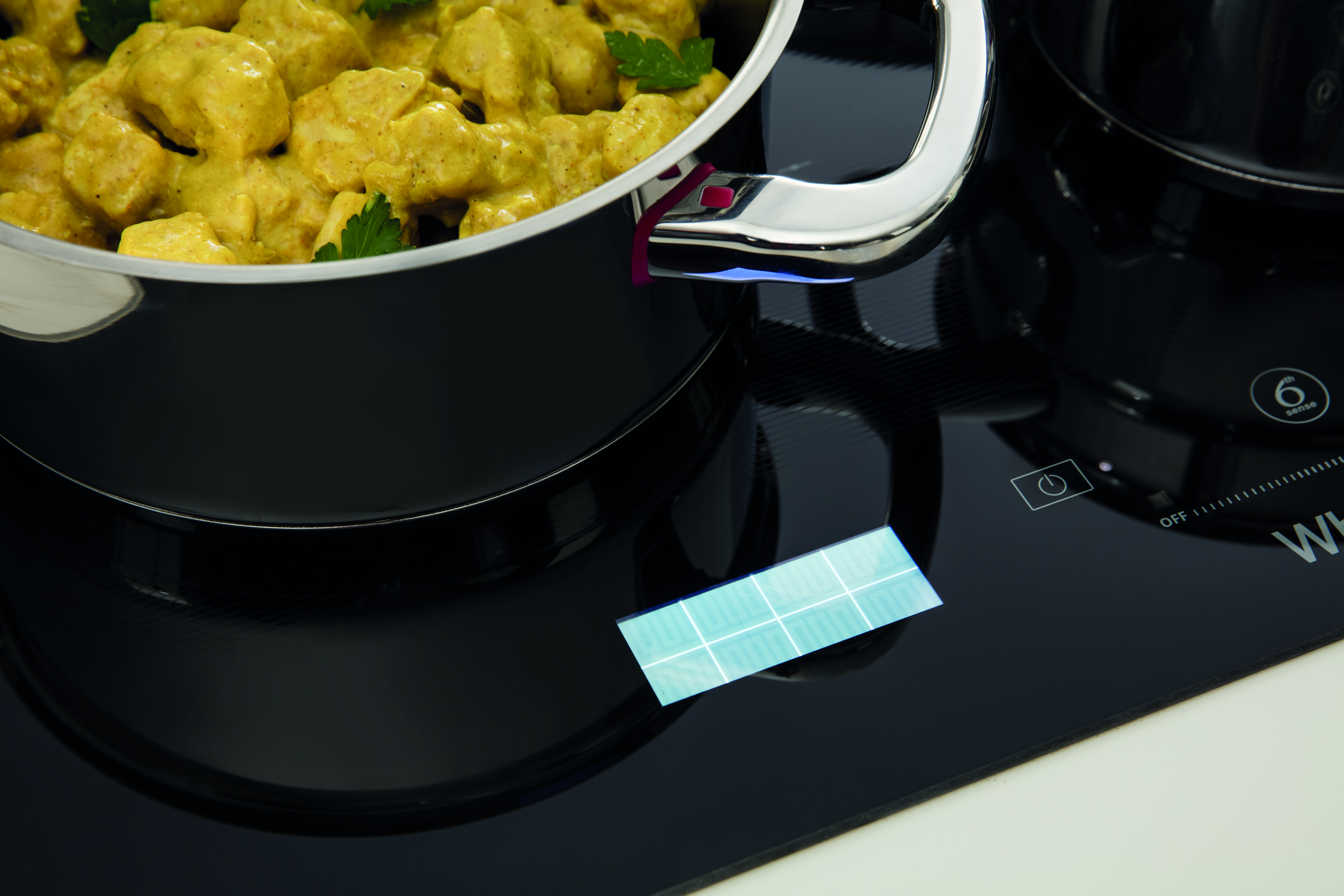 Főzőedény sárga curryvel fekete indukciós főzőlapon, a vezérlőpanel látható.