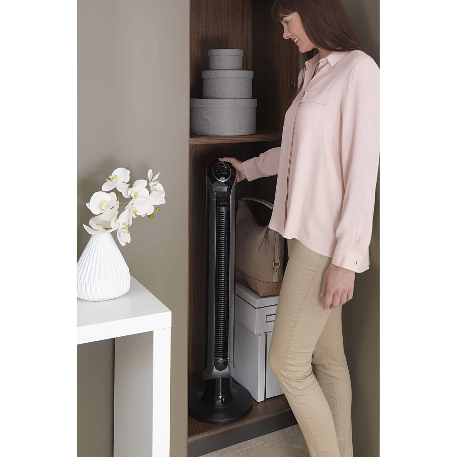 Una donna vicino a un ventilatore nero in un armadio, con un vaso di fiori bianchi sul tavolo.