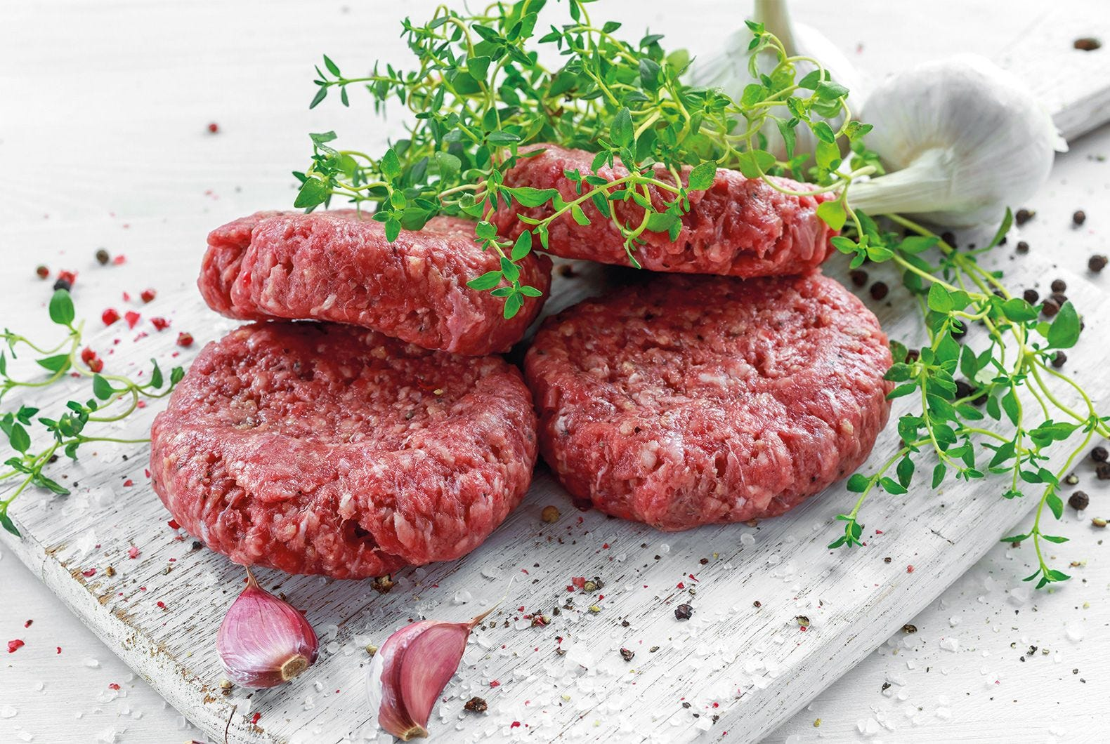 Négy nyers hamburgerpogácsa fehér fa deszkán, fűszernövényekkel, fokhagymagerezdekkel és borsszemekkel.