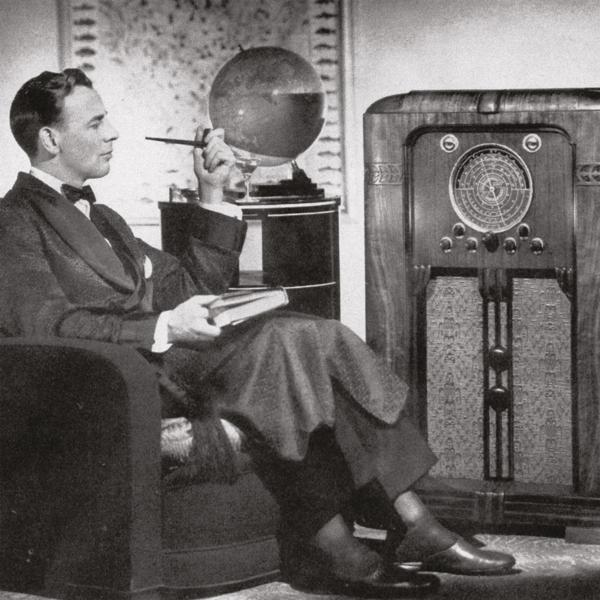 Mann im Anzug sitzt, raucht Pfeife, neben einem Vintage-Radio. Ein Globus steht daneben.