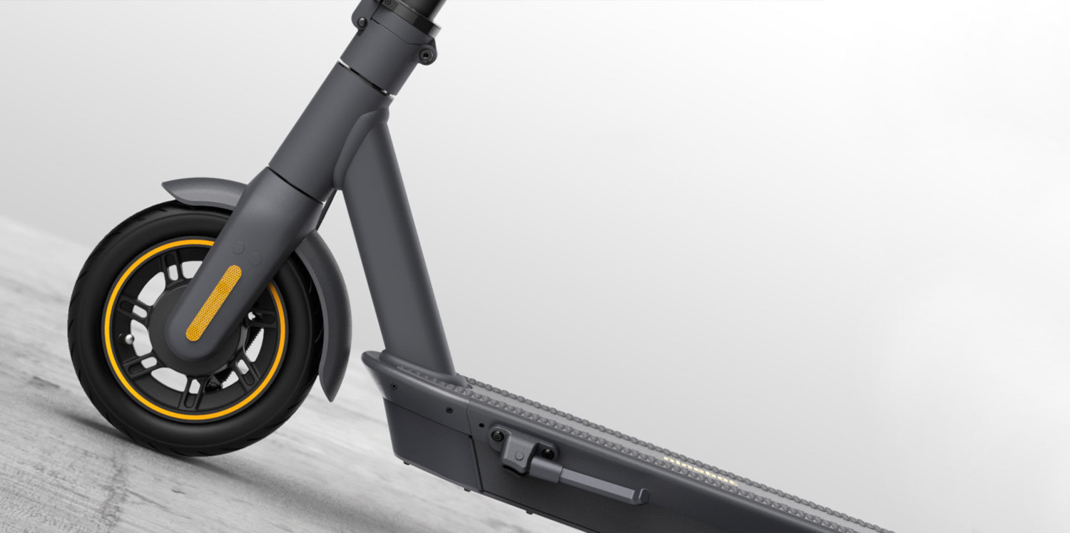 Gros plan d'une trottinette électrique grise. La roue a une jante et un réflecteur jaunes. Le fond est blanc.