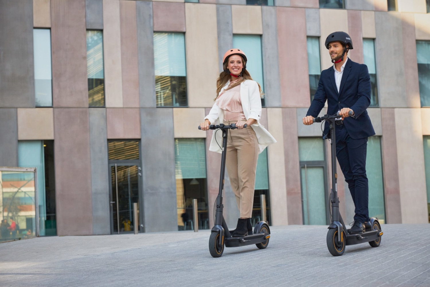 Deux personnes font du scooter devant un bâtiment moderne. Elles portent des casques et des tenues de travail. Jour ensoleillé.