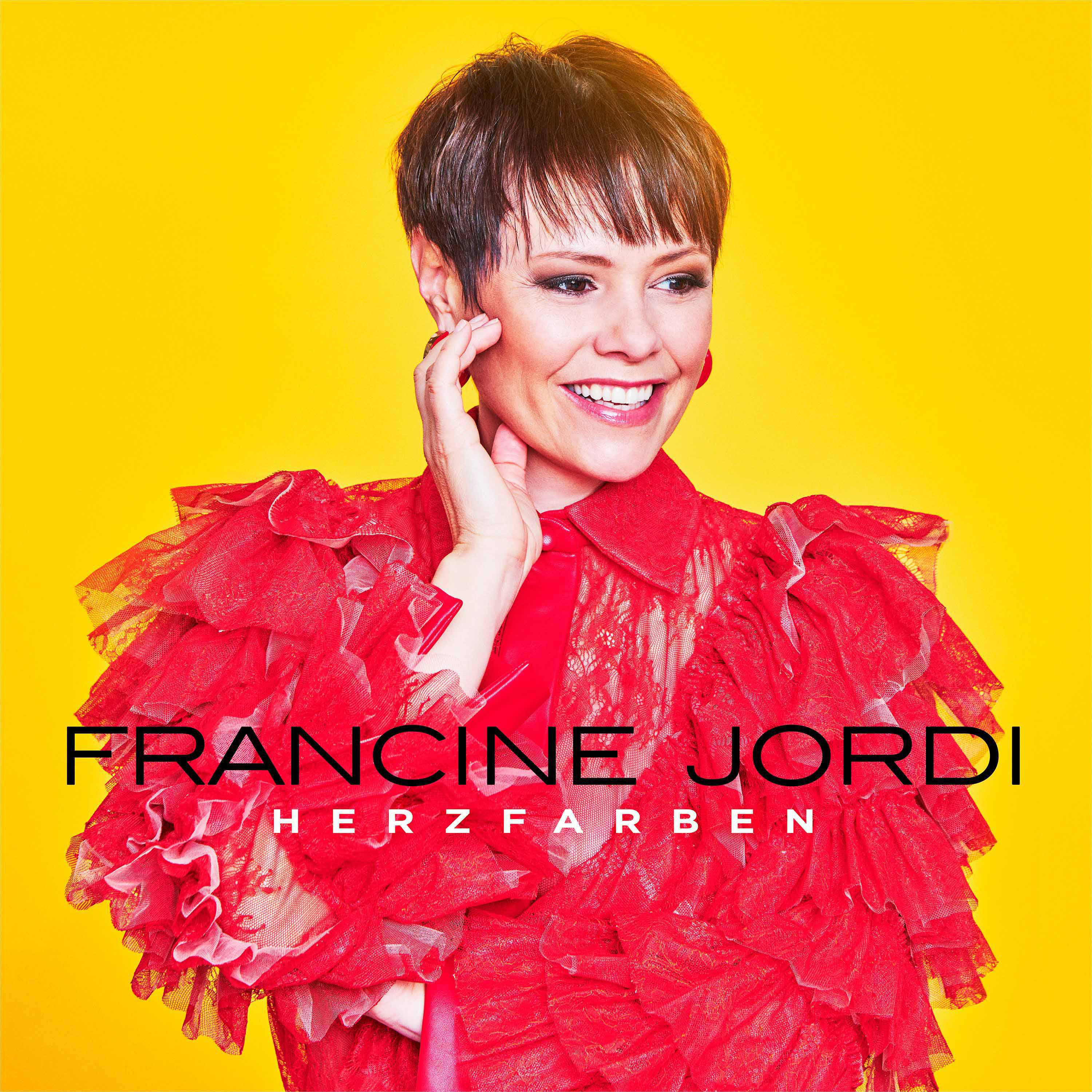 Francine Jordi lächelt, trägt eine rote Rüschenjacke vor gelbem Hintergrund.