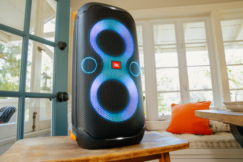 Zwarte luidspreker met blauwe en cyaan lichten op een houten tafel. Een raam en een oranje kussen op de achtergrond.