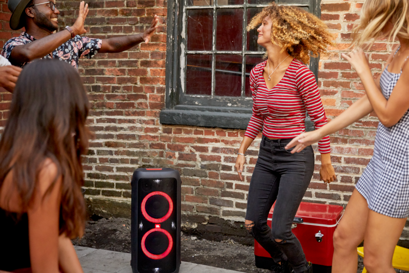 Groep mensen die dansen bij een zwarte luidspreker met rode lichten. Een bakstenen muur en een rode koeler op de achtergrond.