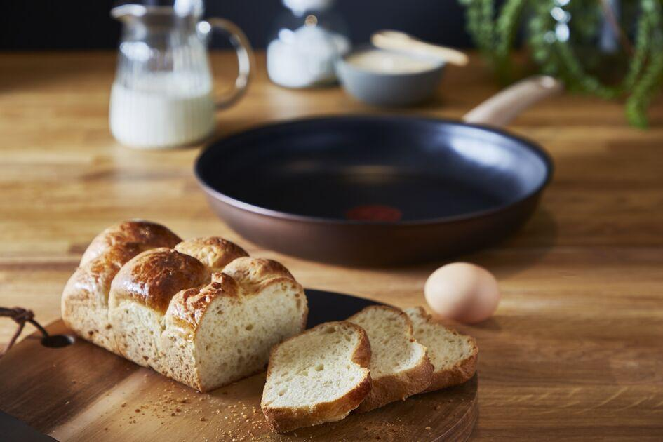 Bread loaf with slices, frying pan, and ingredients on wooden surface. Warm lighting.