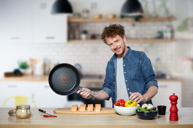 Een man in een spijkeroverhemd houdt een koekenpan vast in een keuken met ingrediënten.