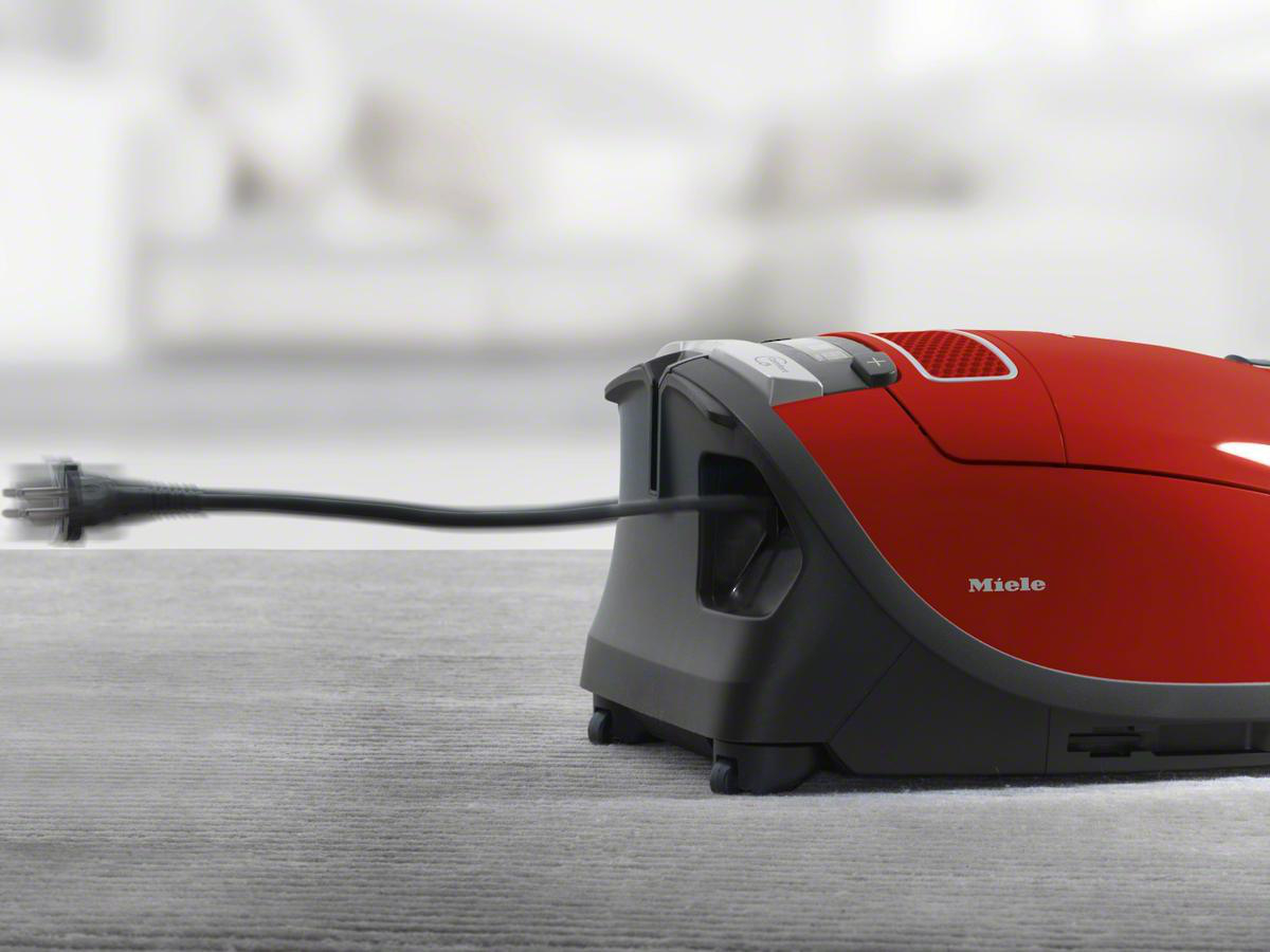 Roter Staubsauger, das Kabel wird eingezogen. Grauer Boden, unscharfer Hintergrund.