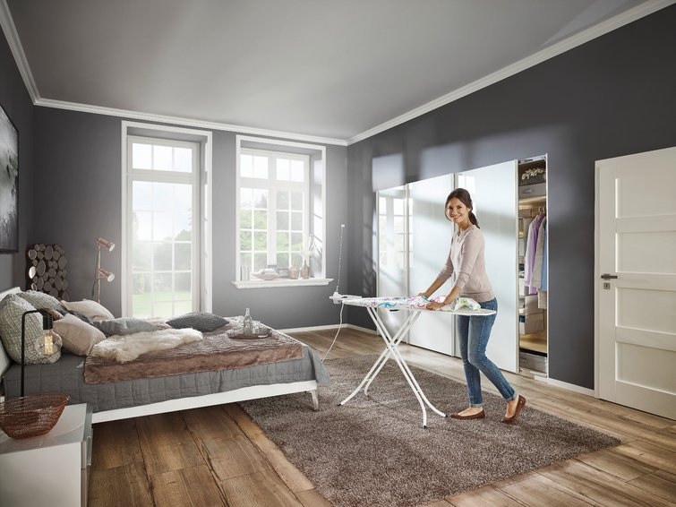 Femme repassant des vêtements dans une chambre. Table à repasser installée. Murs gris.