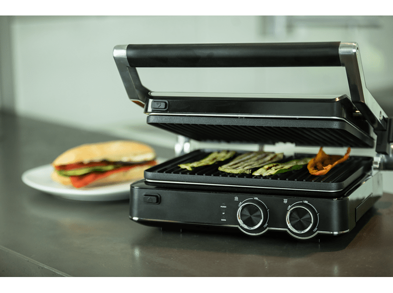Fekete grill nyitott fedéllel, zöldségeket süt. Egy tányér szendviccsel balra.