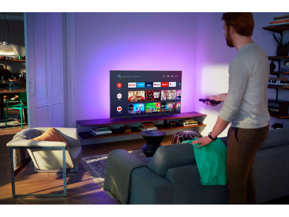 Hombre de pie frente a un televisor inteligente, sosteniendo un control remoto. El televisor muestra una variedad de aplicaciones. Ambiente de sala de estar.