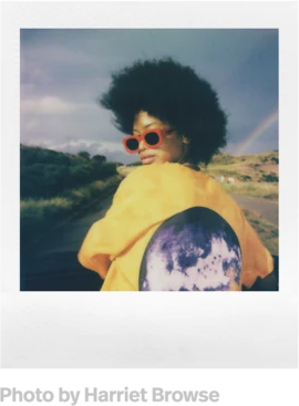 Frau mit Afro, roter Sonnenbrille, gelbem Oberteil und Regenbogen auf einem Polaroid-Foto.