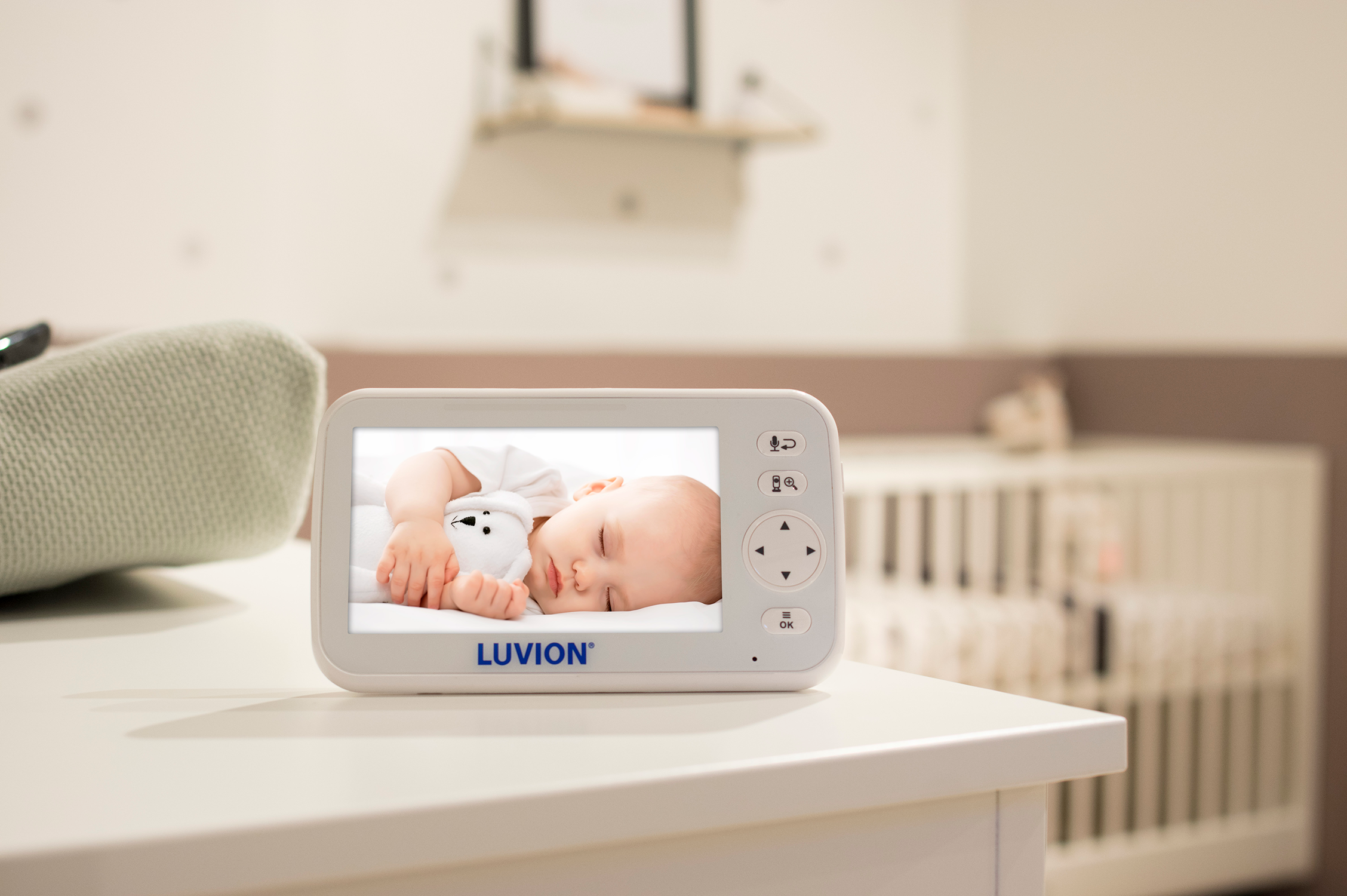 Baby monitor screen displaying a sleeping baby. Monitor is on a white table in a nursery room.