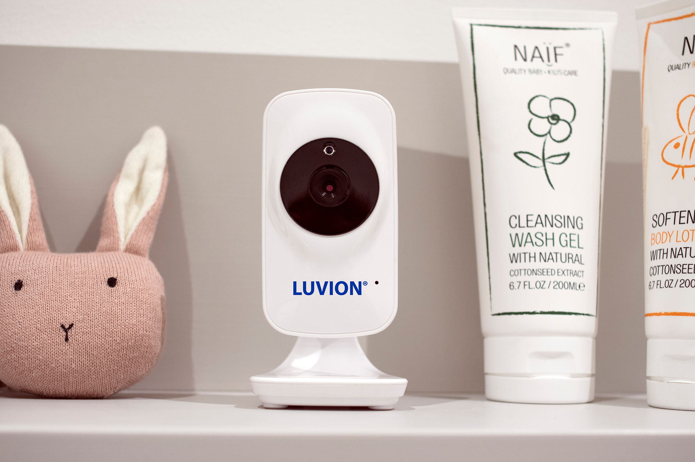 A white baby monitor with a camera and the brand name Luvion. There are baby products in the background.