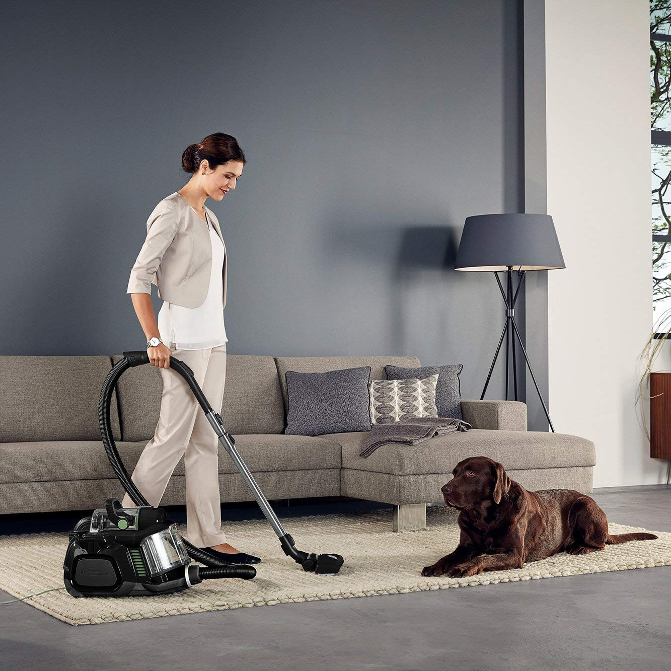 Eine Frau, die einen Teppich in einem Wohnzimmer saugt. Ein brauner Hund liegt auf dem Teppich und schaut zu.