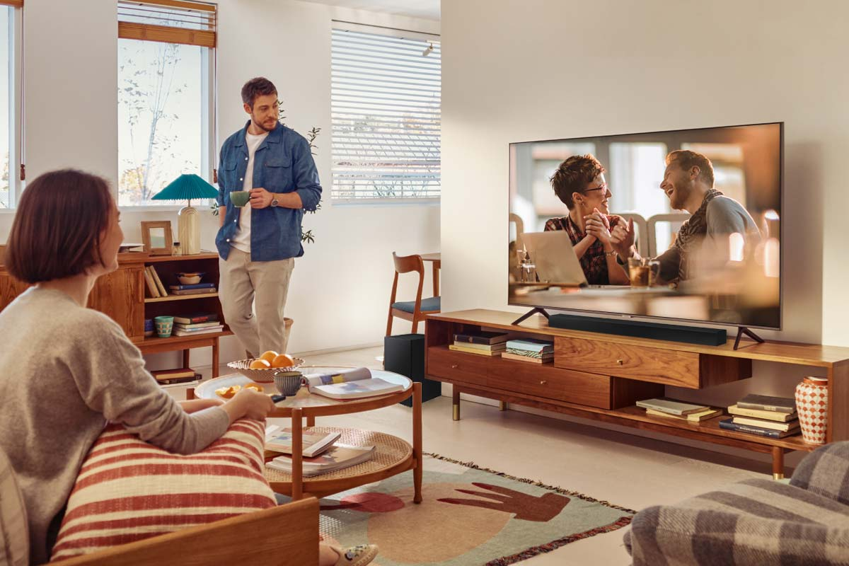 Escena de sala de estar con tres personas. TV muestra a dos personas. Un hombre y una mujer hablan.