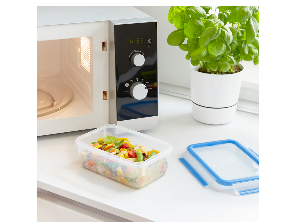 Microondas con recipiente de comida y planta sobre superficie blanca.