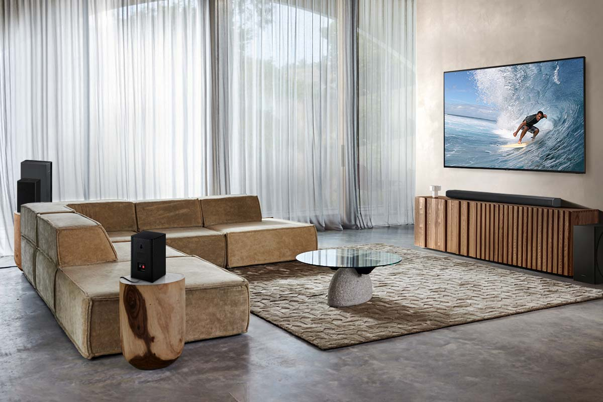 Sala de estar con una gran pantalla de TV que muestra a un surfista. Sofá beige, alfombra, altavoces y muebles de madera.