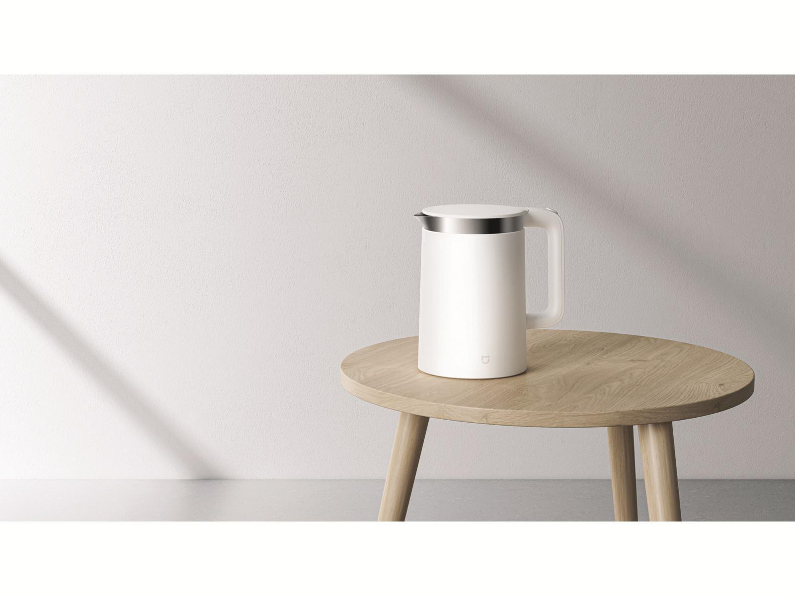 Ein weißer Wasserkocher auf einem Holztisch vor einer weißen Wand. Sonnenlicht ist sichtbar.